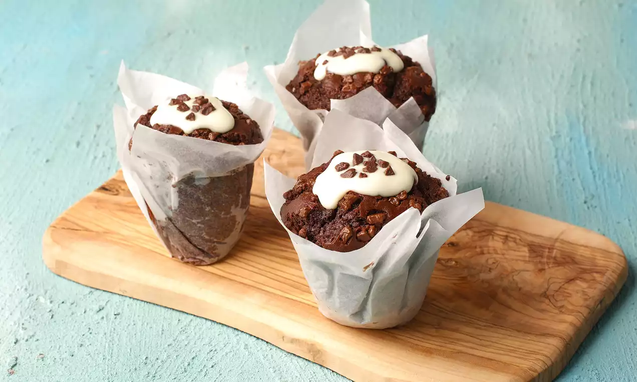 Kenarları yağlı kağıtla sarılmış, üstü beyaz krema ve çikolata parçalarıyla süslenmiş, ahşap bir sunum tahtasında üç çikolatalı muffin