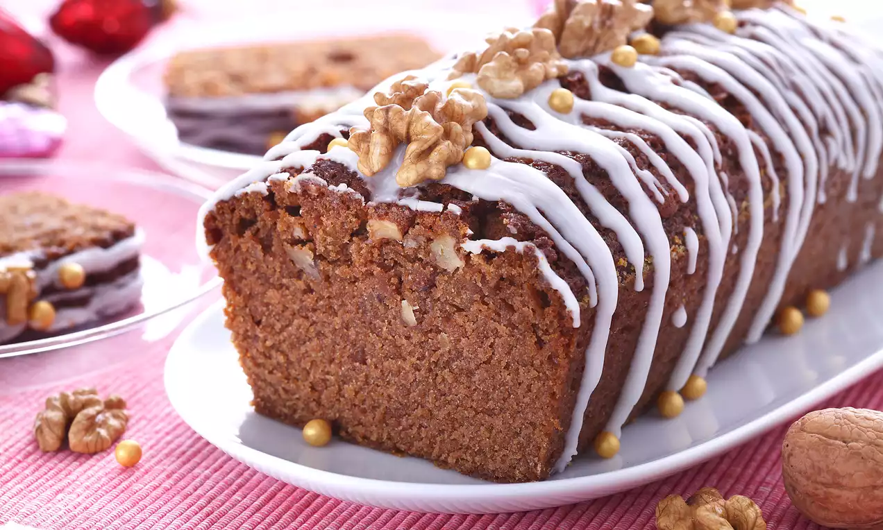 Piernik w formie ciasta ucieranego z widocznymi orzechami włoskimi, polany białym lukrem, ozdobiony złotymi kuleczkami na białej paterze