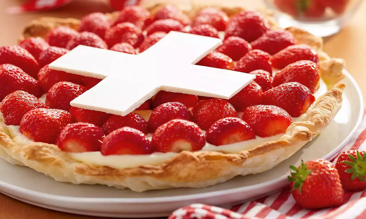 Torta con base di pasta dorata croccante, crema chiara e fragole rosse lucide, decorata con una croce bianca centrale su piatto bianco