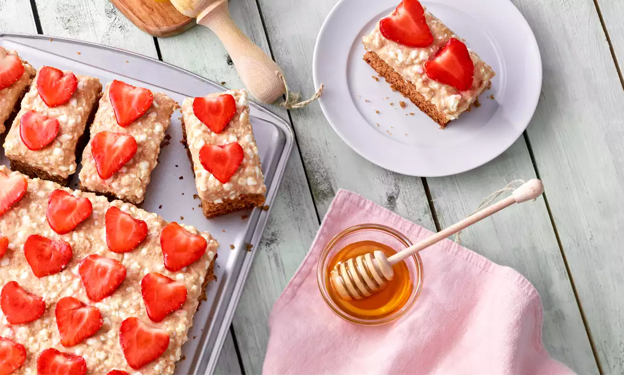 Vollkorn-Kuchen mit hellem Belag und roten, herzförmigen Erdbeerscheiben auf einem Blech neben einem Teller und Honigglas mit Holzlöffel