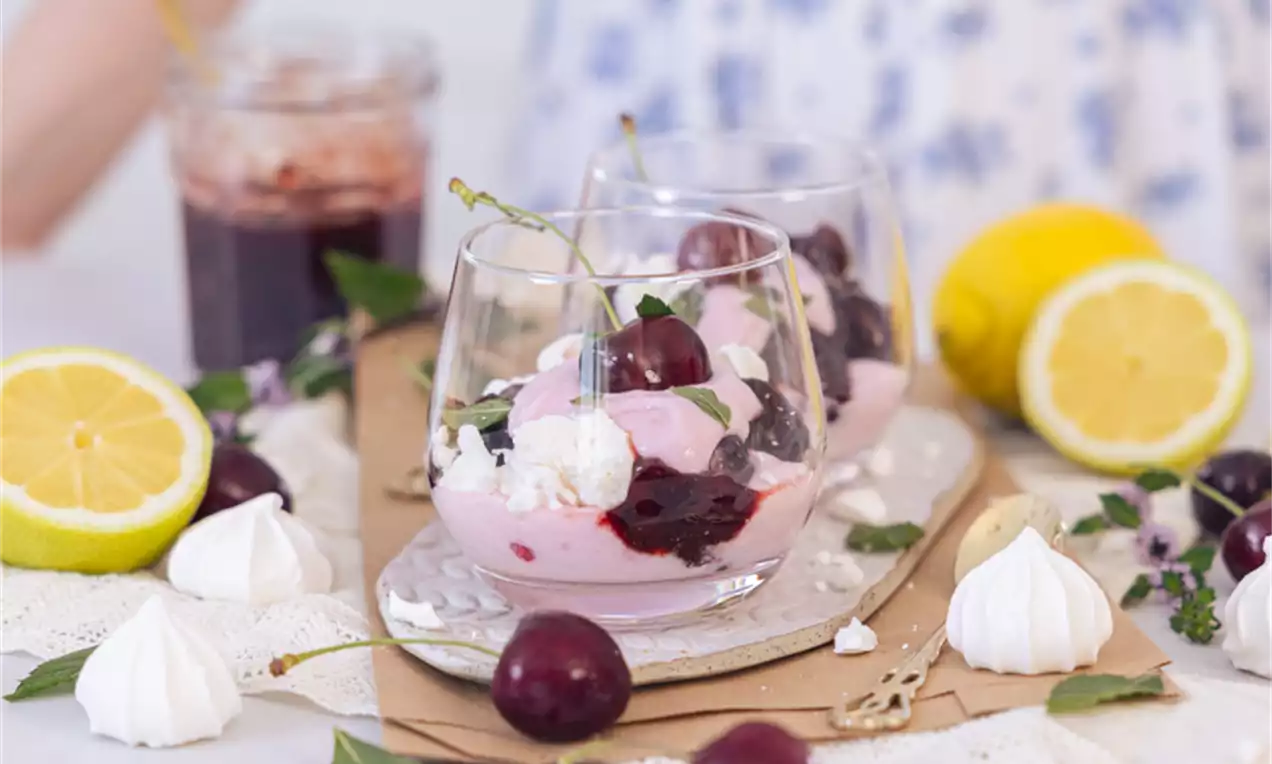 V steklenem kozarcu plast rožnate kreme, temno rdečega češnjevega preliva, belih mereng in svežih češenj na leseni deski z limonami in zelišči