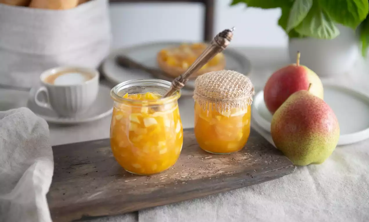 Dva skleněné pohárky plné jasně oranžového džemu s viditelnými kousky ovoce na rustikálním prkénku, se lžičkou a podzimními hruškami