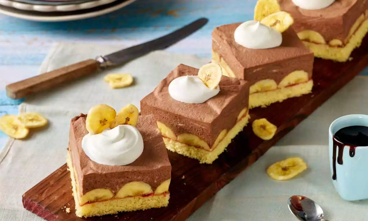 Bananensplit-Schnitten mit gelben Bananenscheiben auf hellem Boden, cremiger Schokoschicht und Sahnetupfen, angerichtet auf dunklem Holzbrett