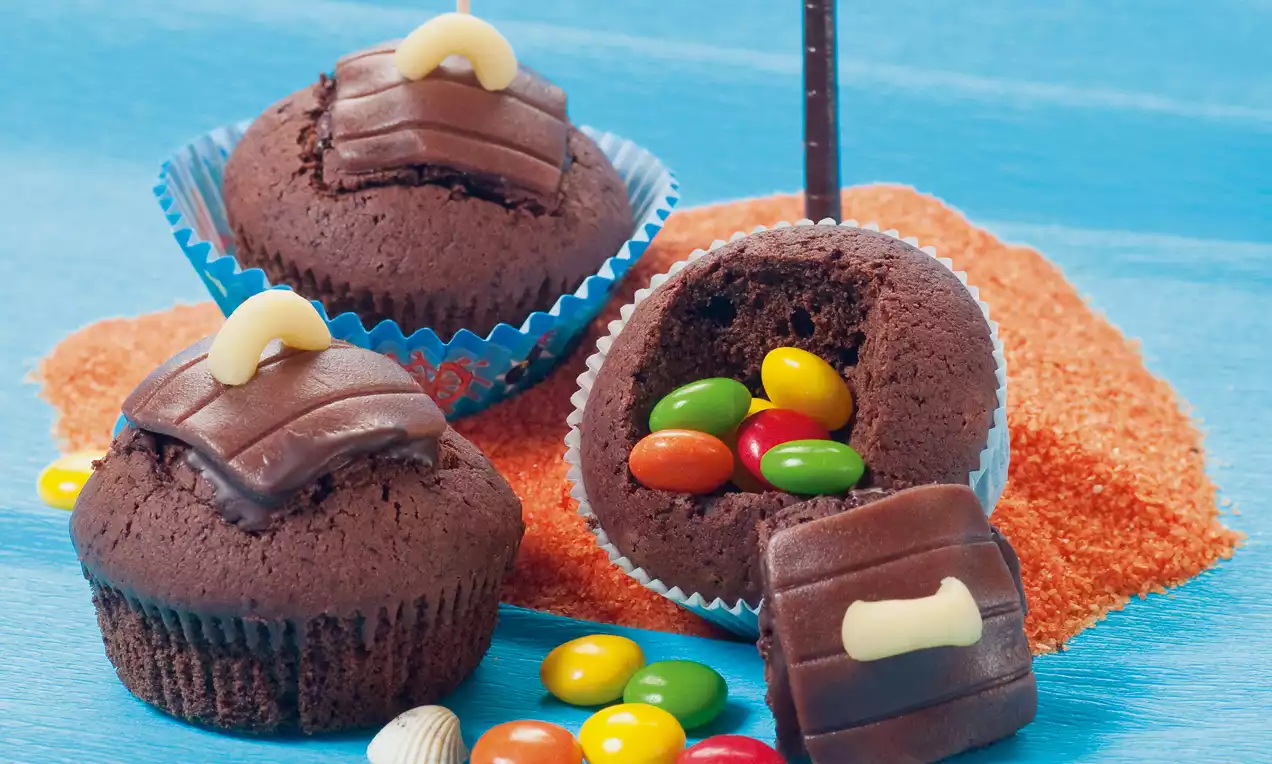Cupcake al cioccolato scuro con copertura lucida e chiusura a forma di scrigno, ripieno di caramelle colorate su fondo azzurro e sabbia arancione