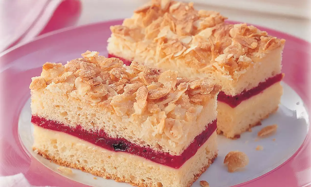 Zwei goldbraune Bienenstichwürfel mit Mandelkruste, heller Cremefüllung und rotem Fruchtgelee auf einem rosa-weißen Teller