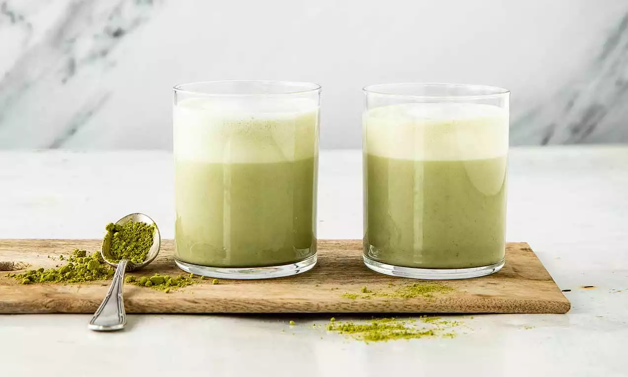 Due bicchieri di latte matcha-vaniglia color verde tenue con strato superiore di schiuma bianca su un tagliere di legno accanto a cucchiaio di polvere di matcha