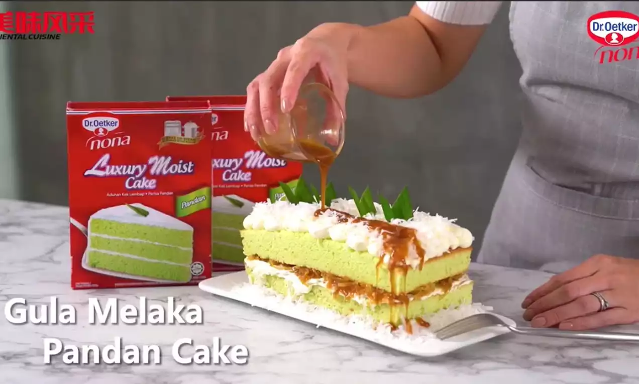 Layered Gula Melaka Pandan Cake with vibrant green sponge, white coconut topping, caramel drizzle and pandan leaves on a white plate