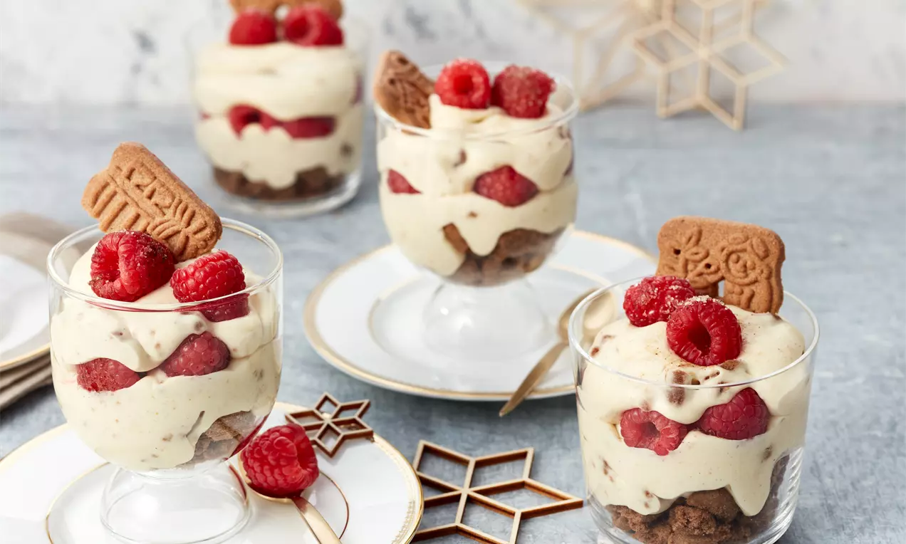 Desert vegan de Crăciun în cupe de sticlă stratificate cu cremă deschisă la culoare, zmeură roșie vibrantă și biscuiți Speculoos crocanți