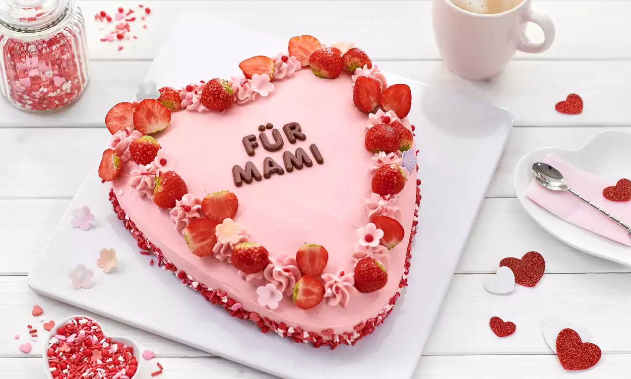 Herzförmige Torte mit zartrosaner Creme, verziert mit Erdbeeren, rosafarbenen Blüten, Schokoladenschrift und roten Streuseln auf weißer Platte