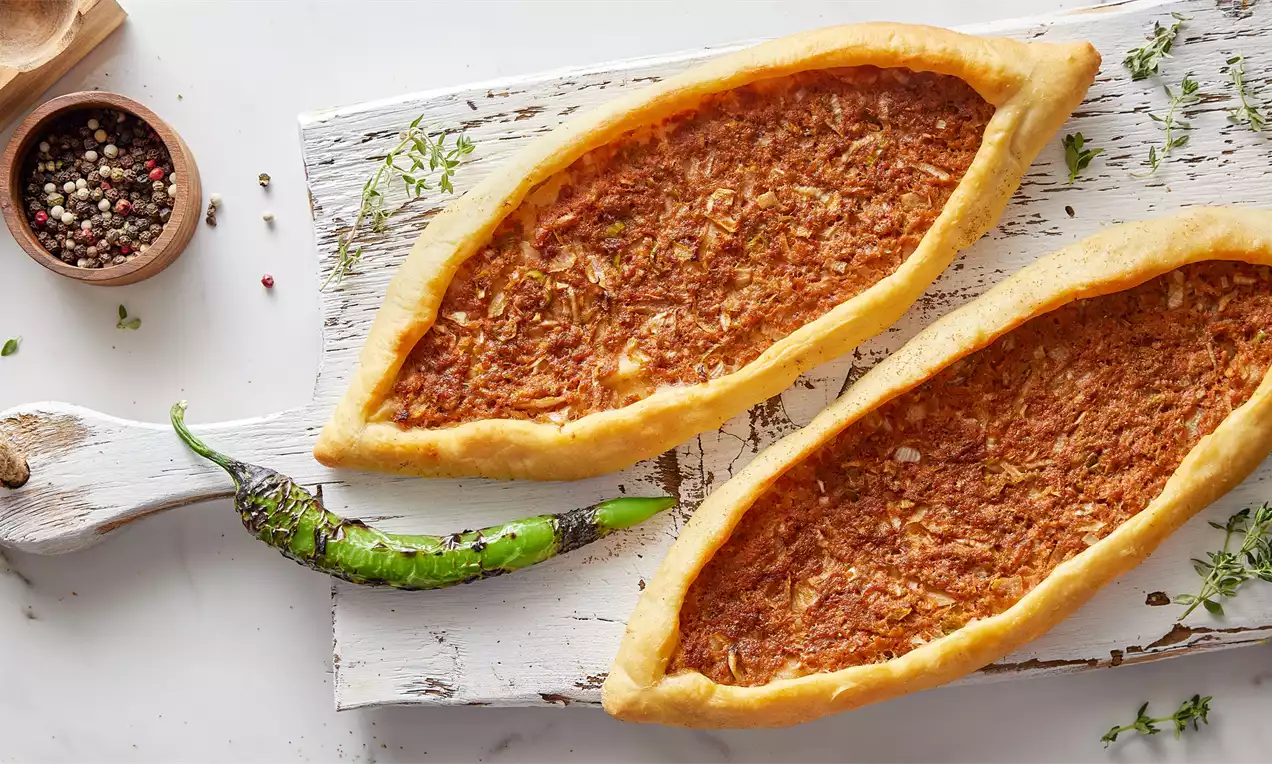 Kayık şeklinde ince ve altın rengi kenarlı kıymalı pideler, beyaz ahşap bir zeminde, yanında yeşil biber ve baharat serpiştirilmiş