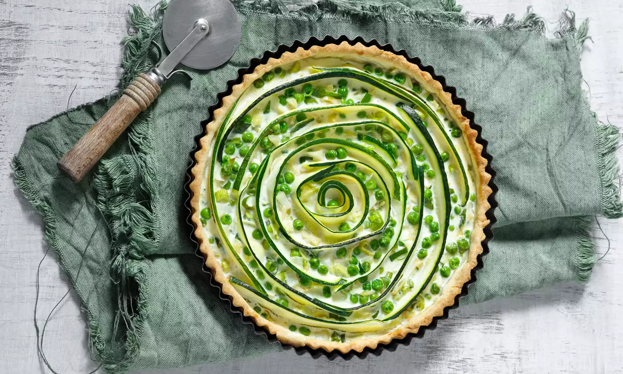 Cuketový koláč s jemne dozlatista upečeným okrajom, zelenou špirálou z tenkých pásikov cukety a hráškom na krémovo bielej plnke