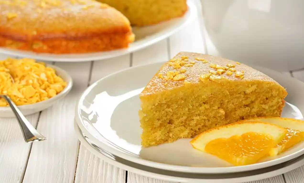 Golden yellow slice of moist oats orange butter cake topped with zest, served on a white plate with a fresh orange slice