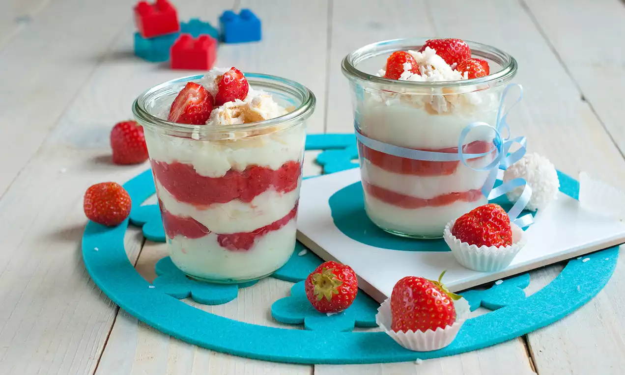 Sklenice s vrstveným kokosovým krémem a jahodovou směsí, zdobené jahodami, drobenkou a kokosovým cukrovím na modré podložce