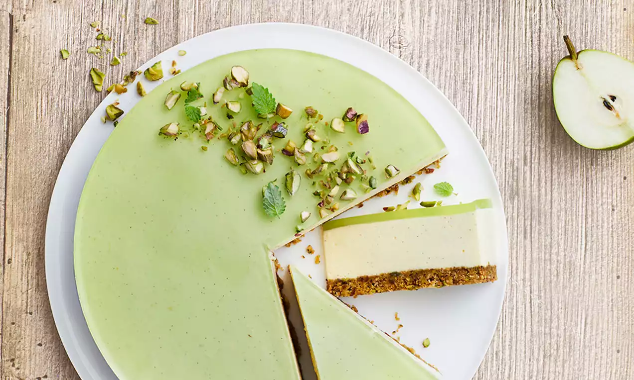 Cheesecake med glat lysegrøn lime-overflade, pyntet med pistacienødder og mynteblade, på sprød brun bund og hvidt fad