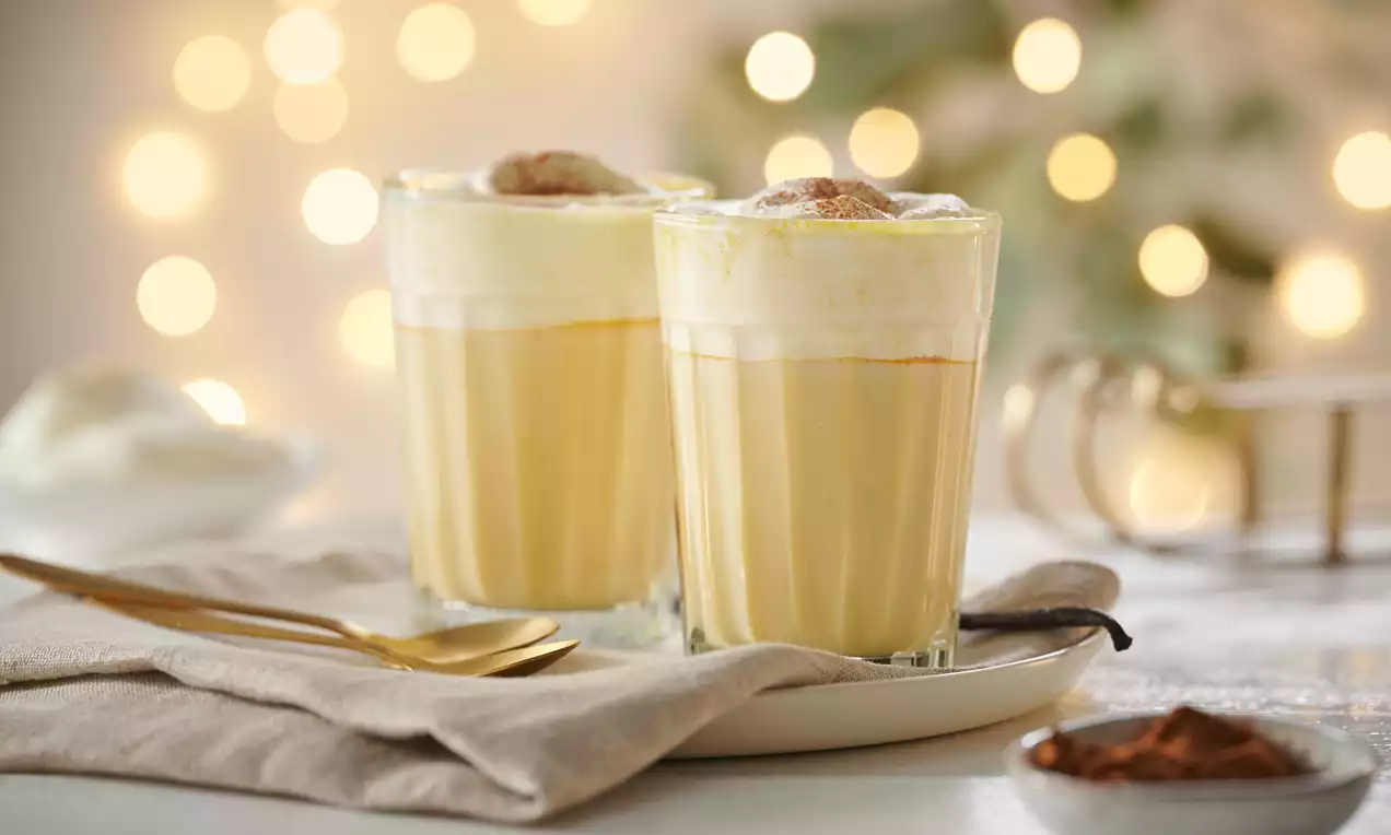 Zwei transparente Gläser mit cremigem gelben Eggnog, bedeckt mit weißem Schaum und Zimt, arrangiert auf einem weißen Teller mit Stoffserviette