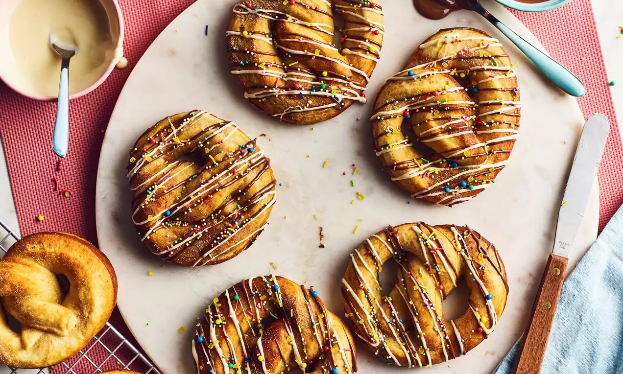 Golden-brown pretzels with a glossy finish and twisted shape arranged on a cooling rack with bowls of creamy chocolate and vanilla sauces nearby