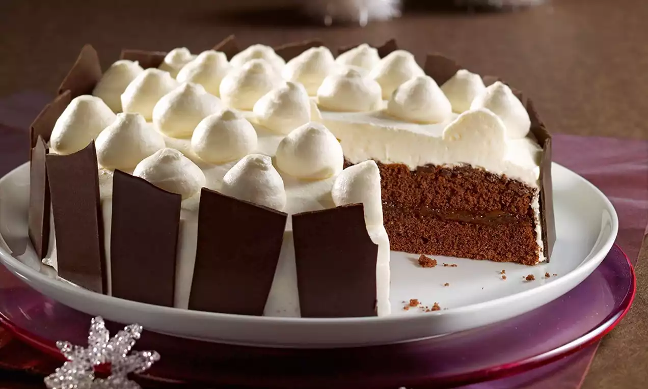 Beyaz kremayla kaplı, tepesinde kabarık krema noktaları olan, kenarları çikolata şeritleriyle süslenmiş, sade tabakta dilimli pasta