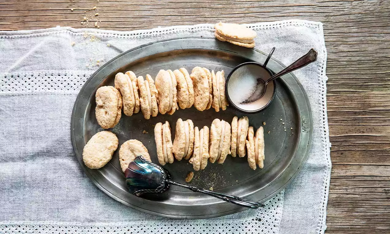 Biscotti ovali dorati accostati su un vassoio metallico, con zucchero in un cucchiaio e sfondo di tessuto leggero su legno