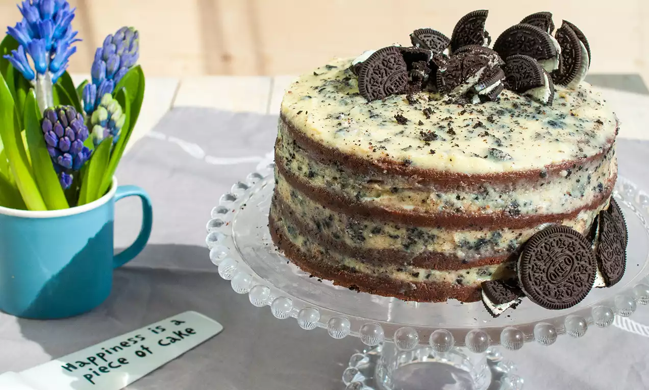 Oreo torta s tmavými čokoládovými vrstvami striedajúcimi svetlý krém, ozdobená kúskami sušienok uložená na sklenenom podnose