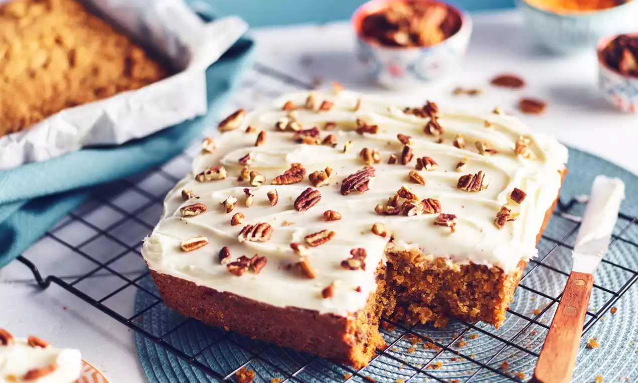 Golden brown carrot tray bake topped with creamy white icing and scattered pecan pieces, resting on a cooling rack with a knife alongside
