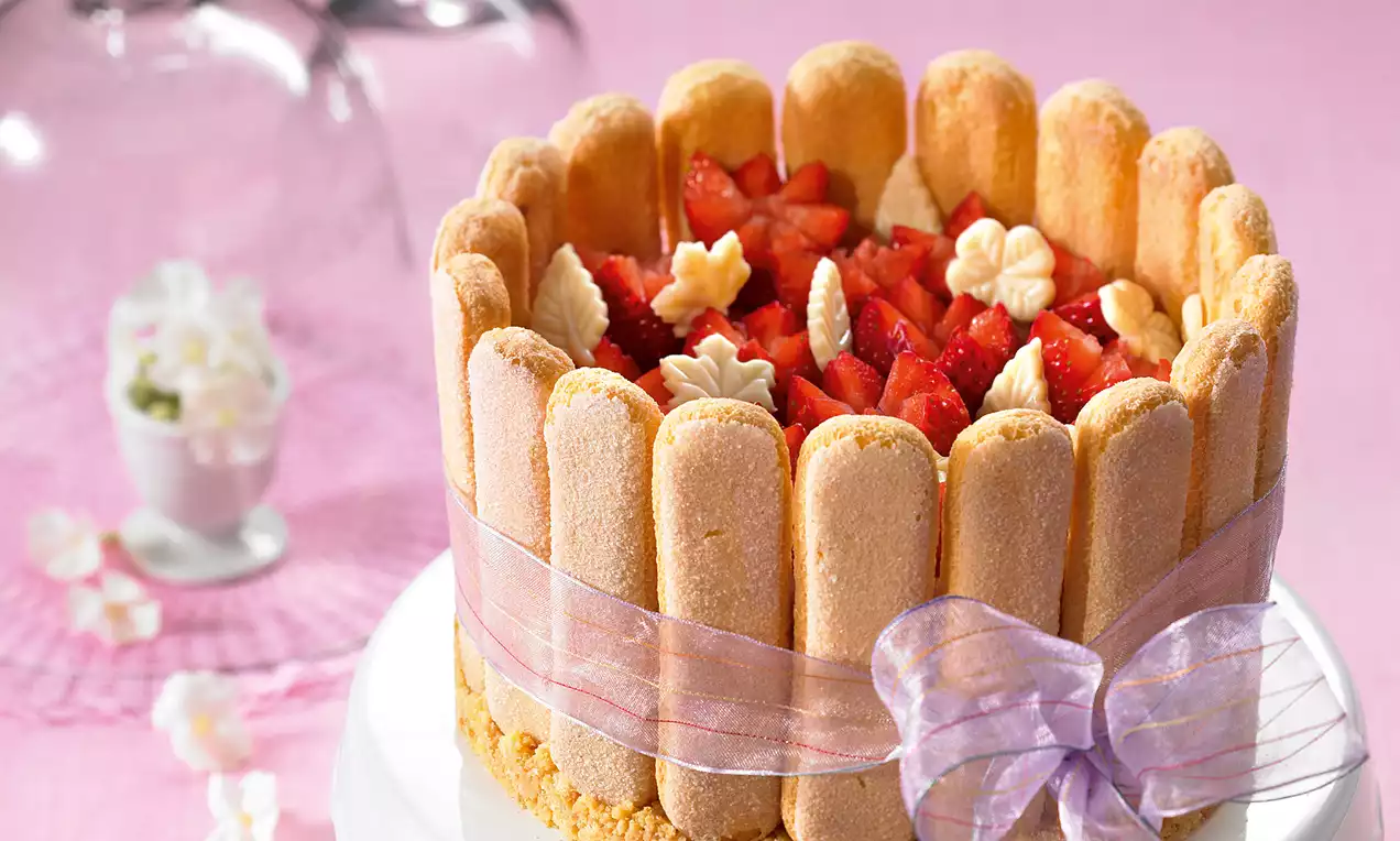 Goldbraune Löffelbiskuits, zu einem Kranz geformt und mit rosa Schleife gebunden, umgeben einen Kern aus Erdbeerstückchen und Sahnetupfen