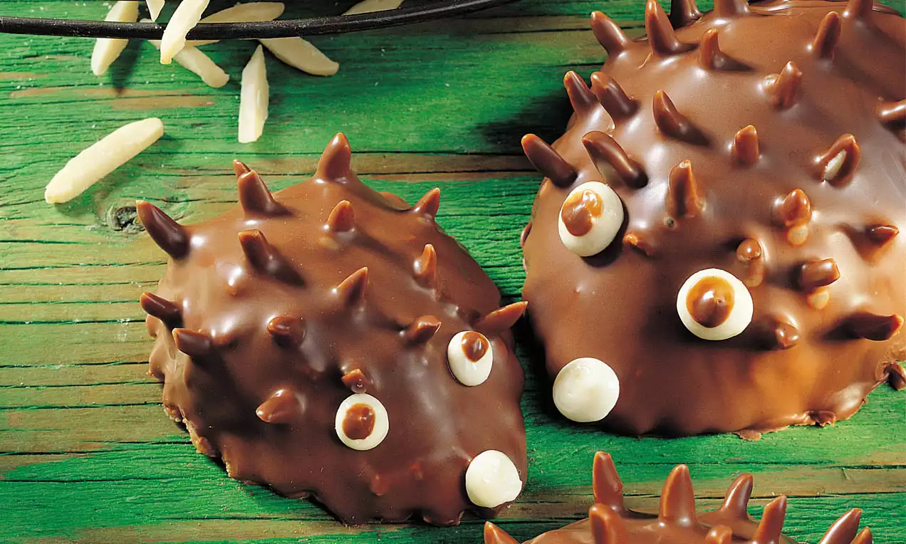 Kleine, braune Schoko-Igel mit spitz zulaufenden Schokoladendornen und weißen Augen auf grünem rustikalem Holzhintergrund mit Mandelstiften