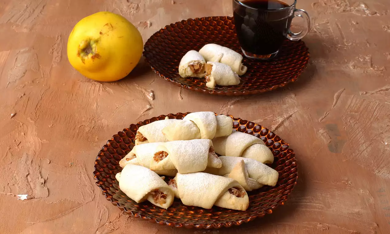 Altın rengi tabağa dizilmiş, ayva dolgulu kıvrık kurabiyeler pudra şekeriyle kaplanmış, yanında bir bardak kahve ve taze ayva yer alıyor