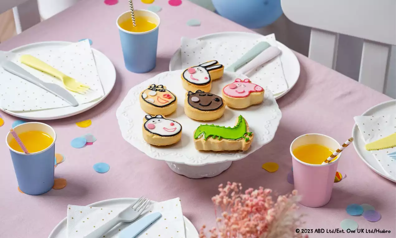 Peppa-Wutz-Kekse mit bunten Gesichtern aus Zuckerglasur und einem grünen Krokodil auf einer weißen Platte, umgeben von Getränkebecher