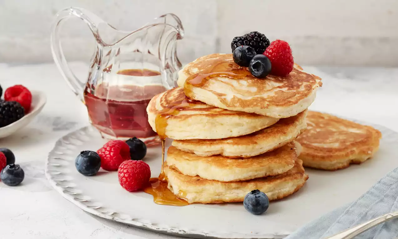 Zlate puhaste palačinke v visokem kupu, prelite z javorjevim sirupom in okrašene s svežimi rdečimi malinami ter črnimi in modrimi borovnicami