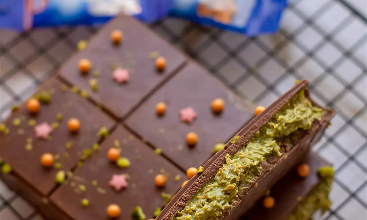 Kvadratići tamne čokolade s glatkom površinom, zelenim slojem pistacija u sredini, ukrašeni šarenim perlicama i zvjezdicama na rešetkastoj podlozi