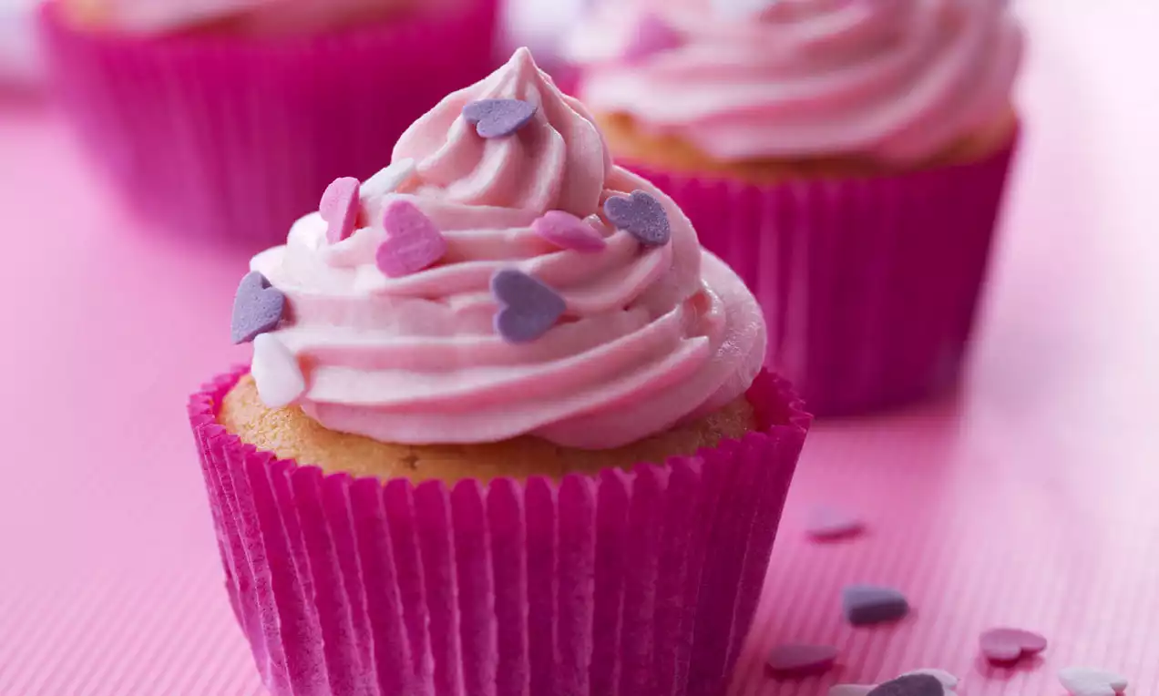 Vaniljecupcake med lyserød flødeskumstop formet som en blød spiral, pyntet med små hjerter og omgivet af en pink papirkapsel