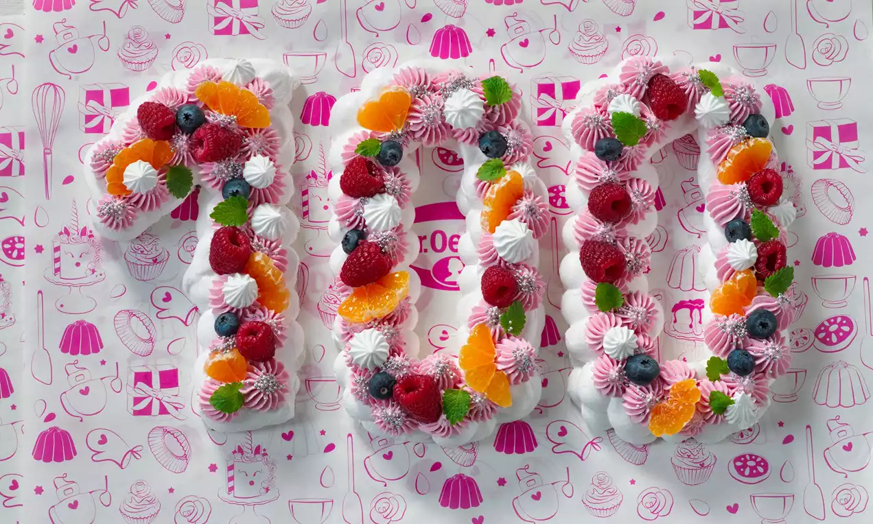 Číslice 100 tvořená bílým a růžovým krémem, zdobená čerstvými malinami, borůvkami, plátky pomeranče a lístky máty na růžovém dekorativním podkladu