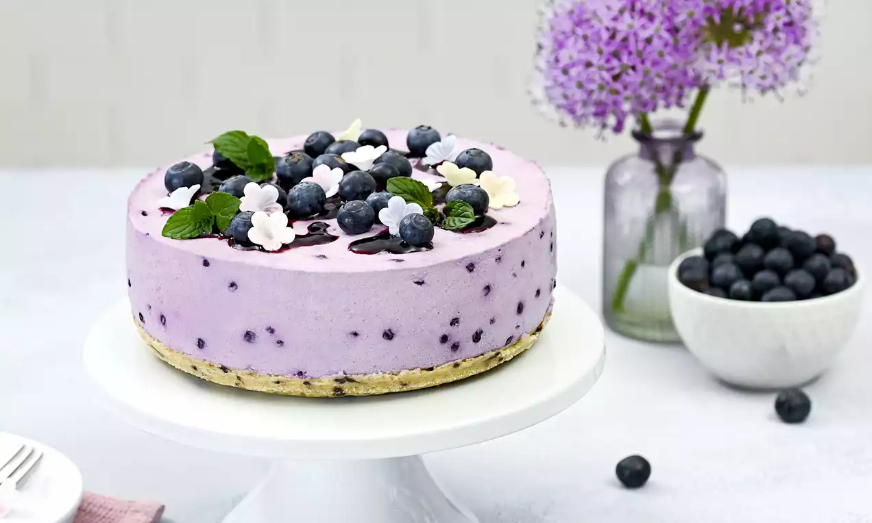 Runder Kuchen mit fliederfarbener Cremefüllung, gesprenkelt mit Blaubeeren auf einem knusprigen Boden, dekoriert mit Blüten und Minzblättern