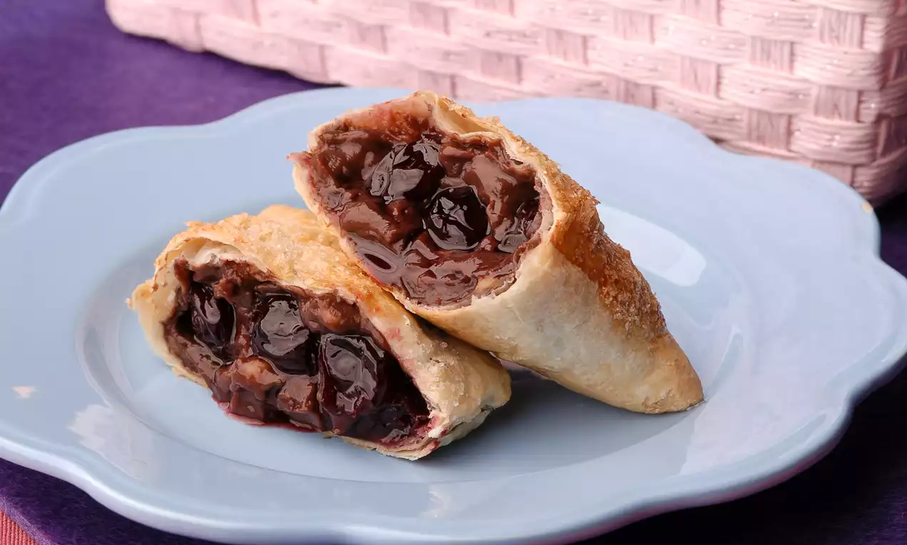 Altın rengi çıtır milföy hamurunda bol vişneli dolgu, katmanları belirgin şekilde ikiye kesilmiş, açık mavi tabakta sunulmuş