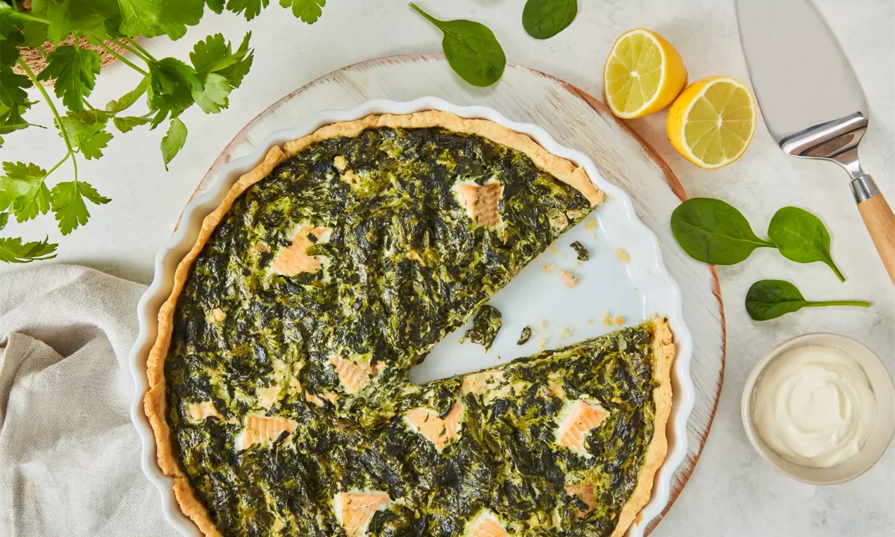 Goldene Quiche mit welligem Rand, grünem Spinatbelag und pastellfarbenen Lachsstückchen in heller Backform, umgeben von frischen Kräutern