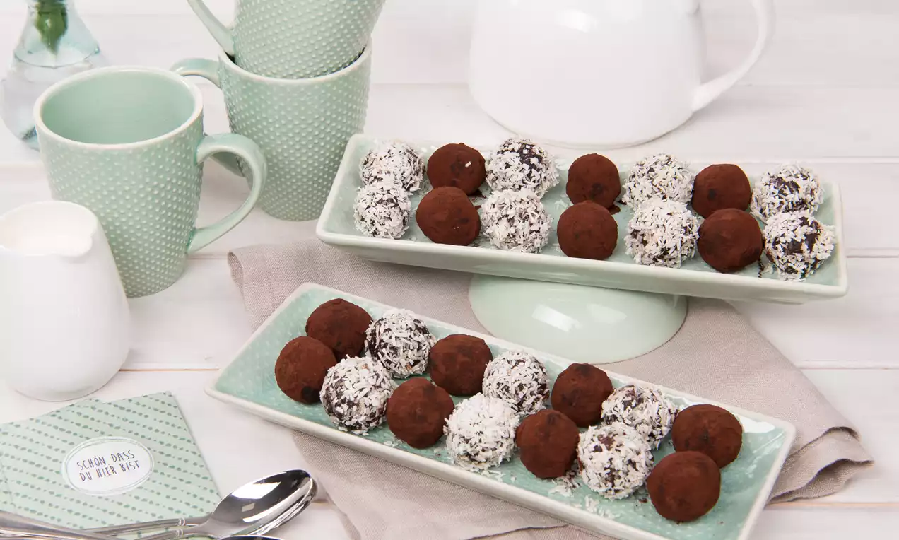 Raw Bites auf hellem Teller, kleine runde Energiebällchen, teils in Kokosraspeln, teils mit Kakaopulver und gehackten Nüssen angerichtet