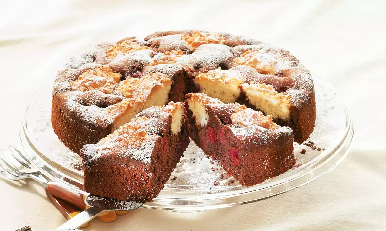 Runder Kirsch-Kokos-Kuchen mit goldbrauner Kruste, roten Kirschstücken, heller Kokosfüllung und Puderzucker auf einer Glasplatte