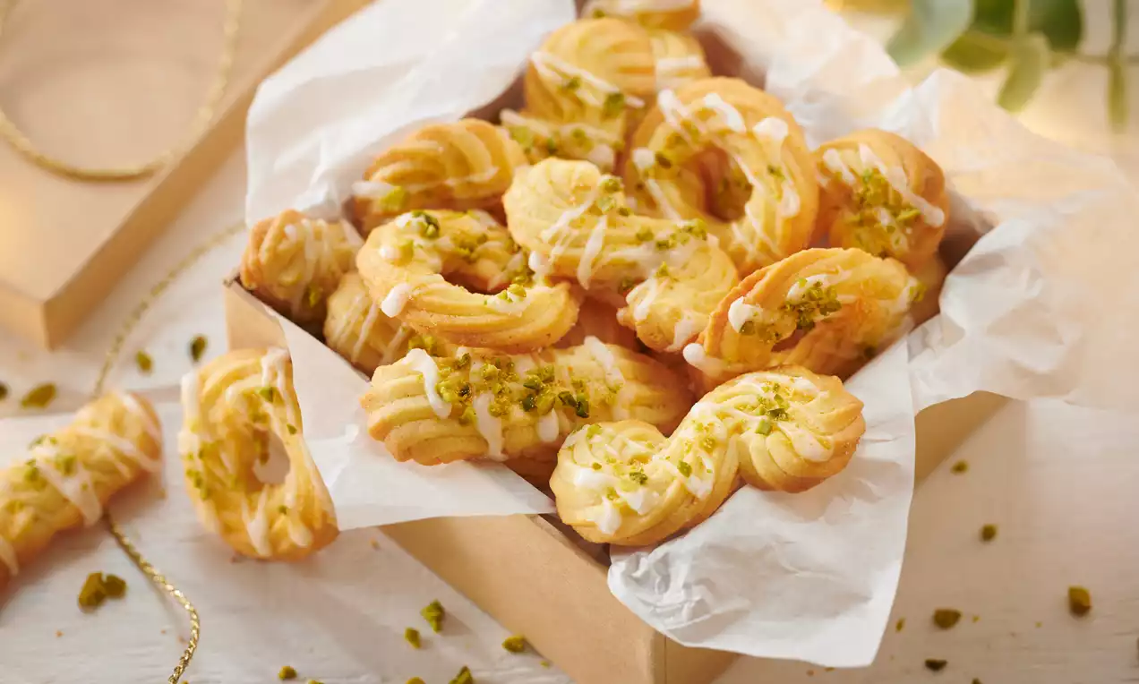 Goldgelbe, spritzförmige Orangenplätzchen mit weißem Zuckerguss und grünen Pistazienstückchen in einer Schachtel auf weißem Backpapier