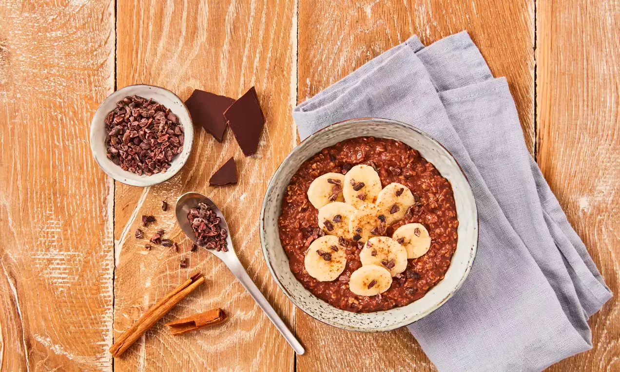 Schokoladenbraunes Porridge mit cremiger Textur, belegt mit frischen Bananenscheiben und Kakaosplittern in einer hellen Schale auf Holzuntergrund