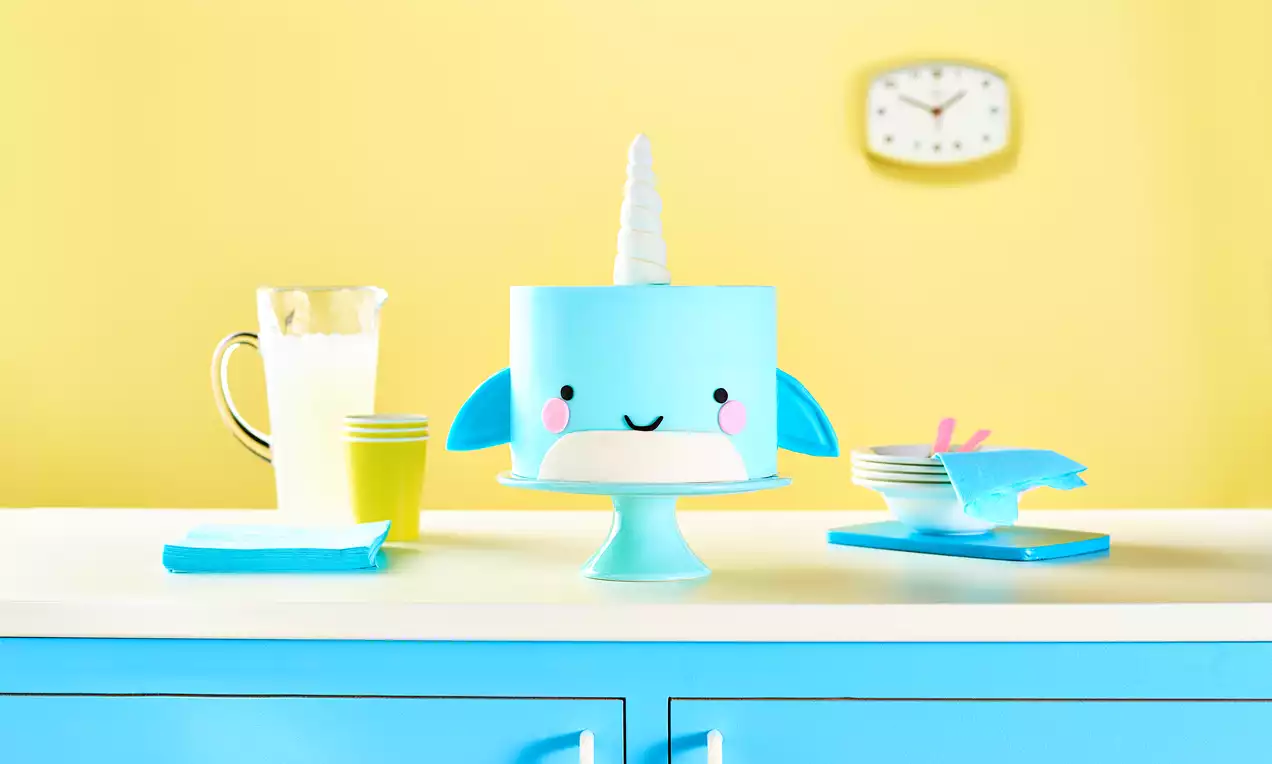 A pale blue round cake shaped as a cute narwhal with a white horn, pink cheeks, and fins, on a matching stand on a white counter with blue cabinets against a yellow wall