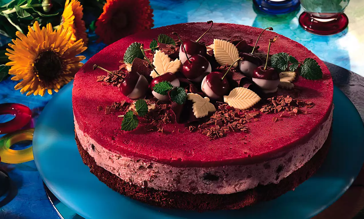 Kirsch-Sahne-Torte mit roter Gelatinehaube, Schokoraspeln, kandierten Kirschen, weißer Schokolade und Minzblättern auf blauem Teller