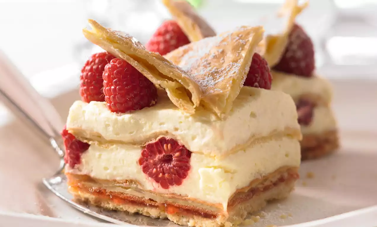 Malinovo-krémový zákusok s vrstvami zlatistého lístkového cesta, žltkastým krémom, červenými malinami a posypom jemného práškového cukru  