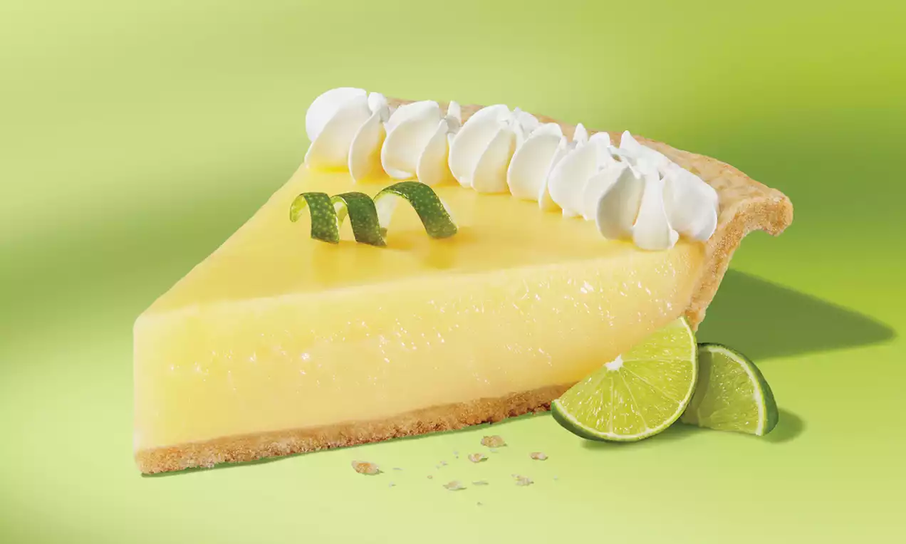 Tranche de tarte Key Lime jaune pâle brillamment lisse sur une croûte dorée, garnie de crème blanche et de zestes de citron vert