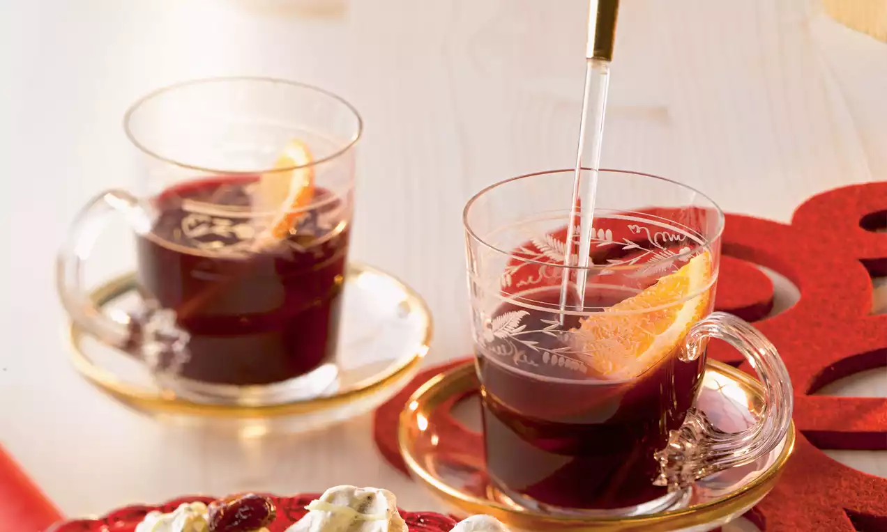 İki ince cam bardakta koyu kırmızı sıcak içecek, içlerinde portakal dilimi ve cam karıştırıcı, altın detaylı fincan tabakları üzerinde 