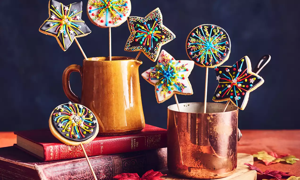 Round and star-shaped Firework Biscuit Pops with vibrant icing patterns and sprinkles, displayed in copper and ceramic pots on books
