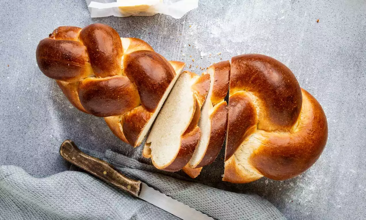 Lesklý zlatohnědý cop z kynutého těsta s měkkou světlou strukturou, částečně nakrájený, položený na šedém povrchu vedle nože