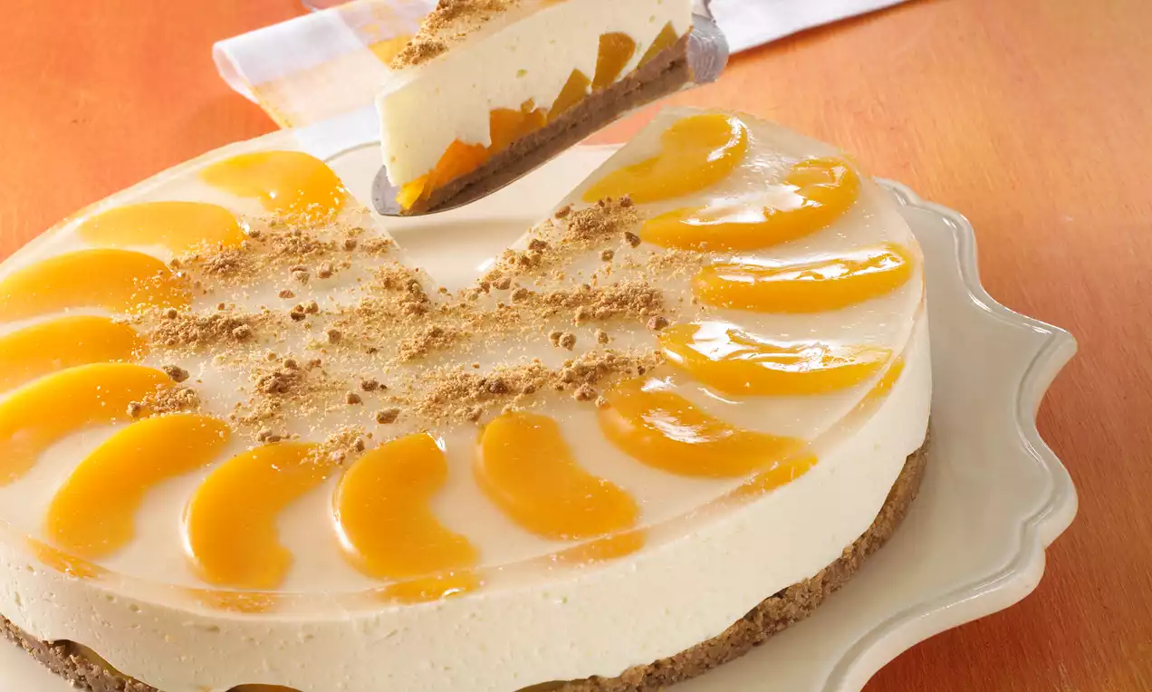 Pfirsch-Sekt-Torte mit hellem Keksboden, cremiger Füllung, glänzenden Pfirsichspalten und Bröseln auf einer weißen Tortenplatte