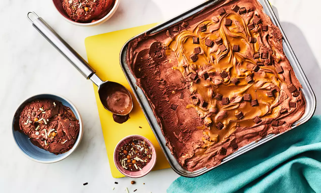 Rich chocolate ice cream swirled with golden caramel, topped with chocolate chunks in a metal tin, served with scoops in small bowls