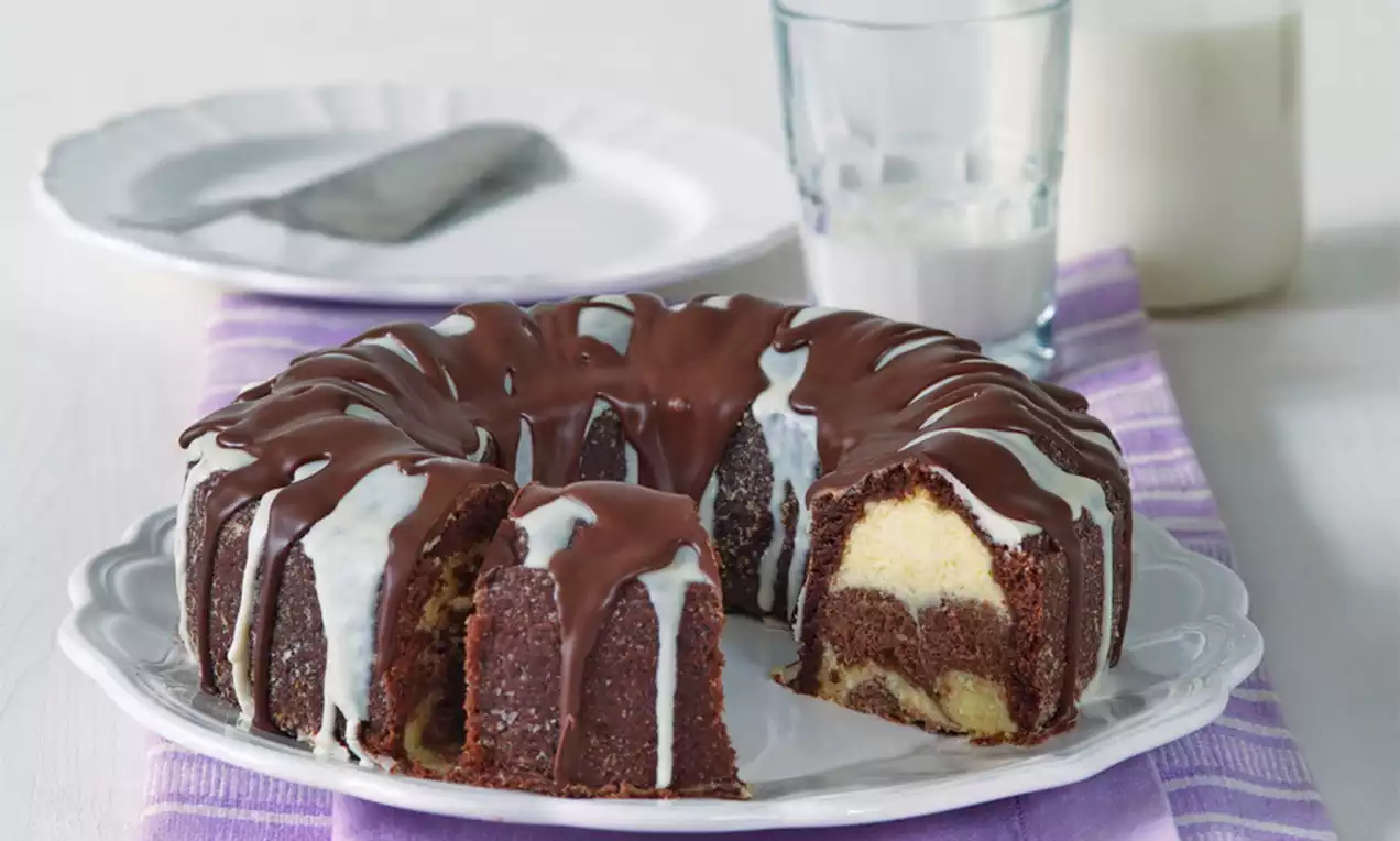 Čokoládovo-tvarohový veniec s mramorovým vzorom, poliaty tmavou a bielou polevou, na bielom tanieri a fialovej utierke s pohármi mlieka