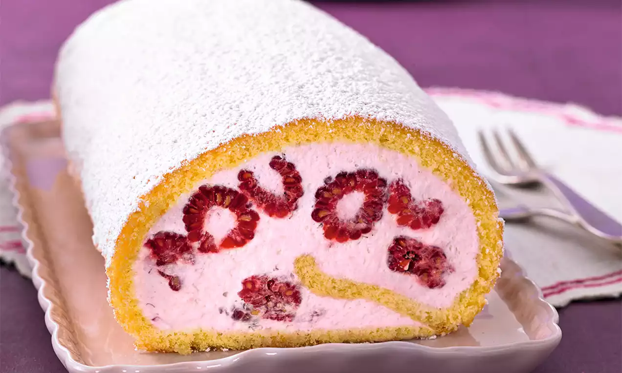 Světle žlutý piškot stočený do rolády s náplní z růžové šlehačky, celými malinami a jemným bílým moučkovým cukrem na růžovém talíři